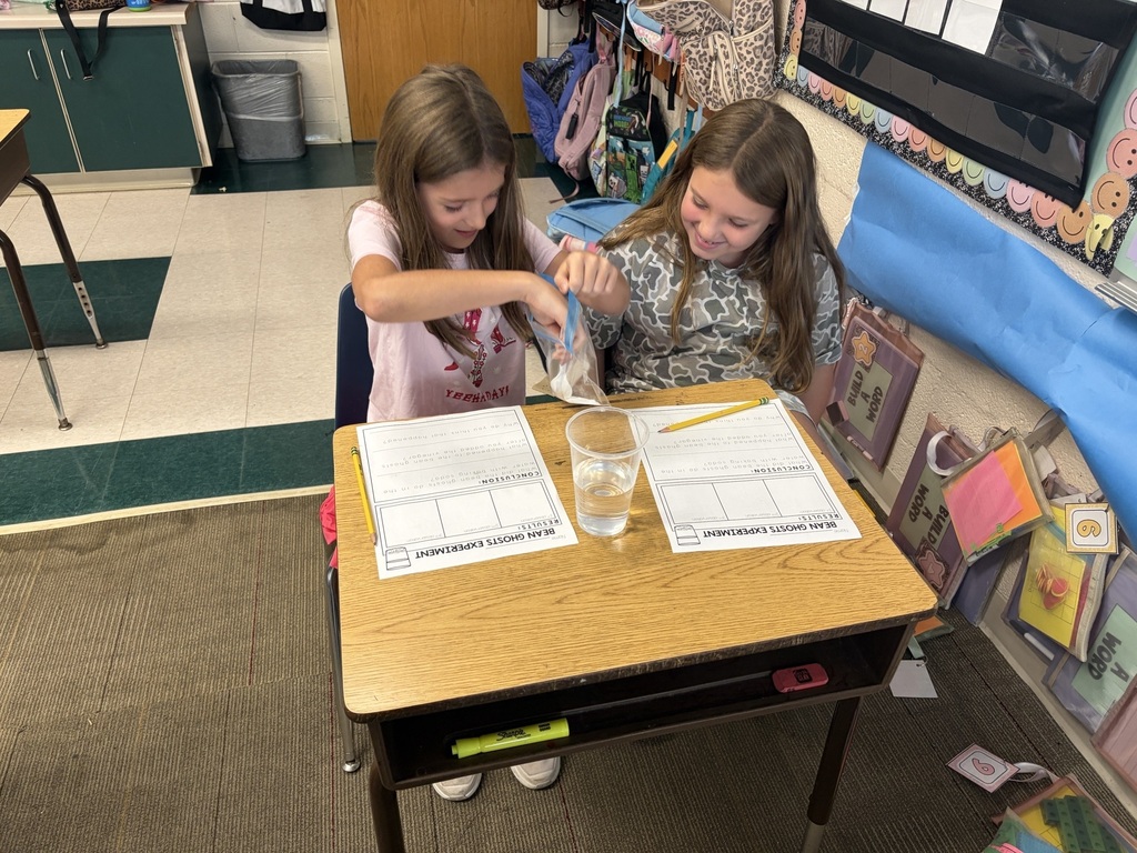 Two girls seated at a desk, one holding a glass of water and the other a small item.