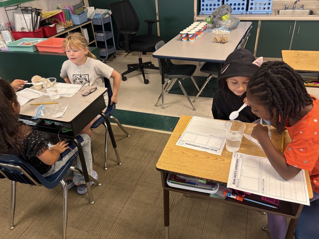 Four children in a classroom with desks, sitting and using spoons to stir contents in clear cups.