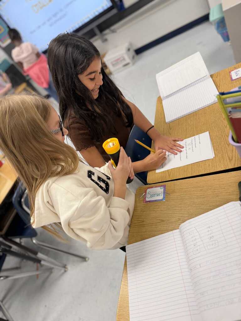 Two girls seated at a desk, one holding a flashlight, another writing on paper.