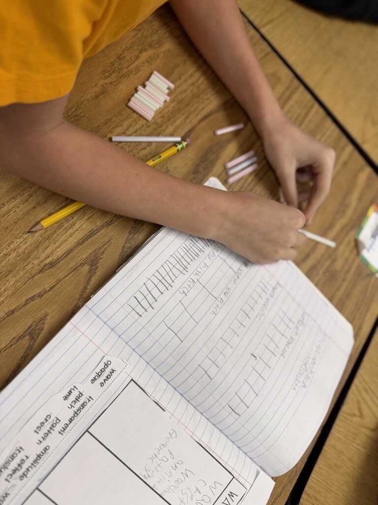 A student sits at a desk, working with small sticks, pencils, and an open notebook.