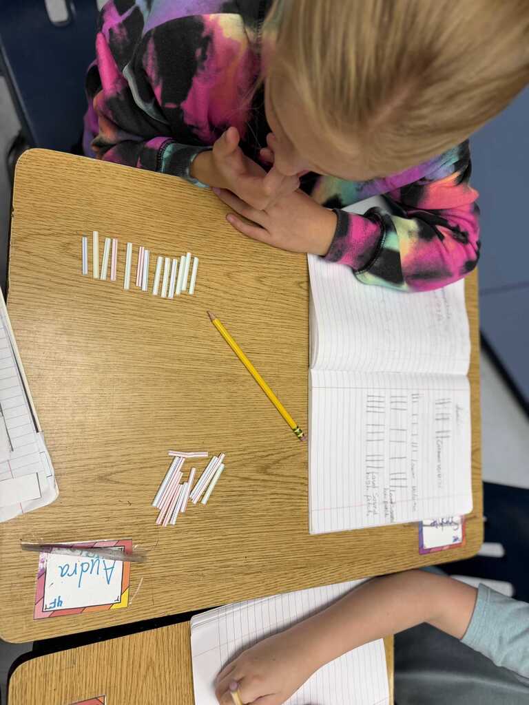 Two children seated at desks with papers and pencils, one arranging small items.