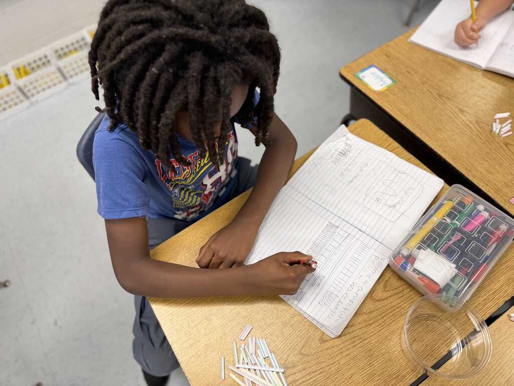 Child seated at desk, writing on paper and using a pencil. Several colored pencils in container on desk.