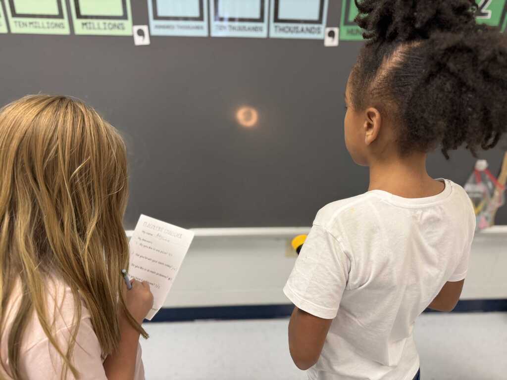 Two children at a school wall, one holding paper, another looking at a red dot on the board.