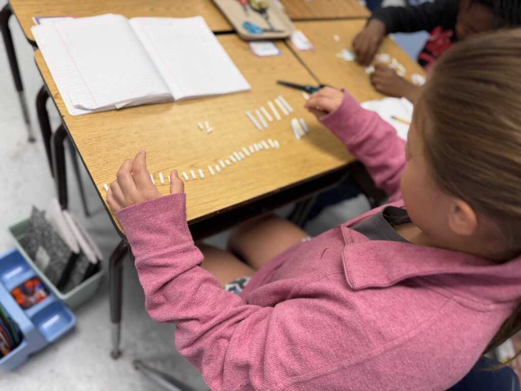 Child seated at a desk, writing on paper with a pen; other student nearby also writing.