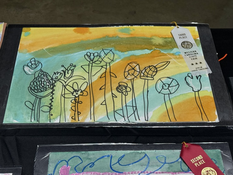 Art work with flowers and a yellow sky, displayed in a black box, with a white tag.