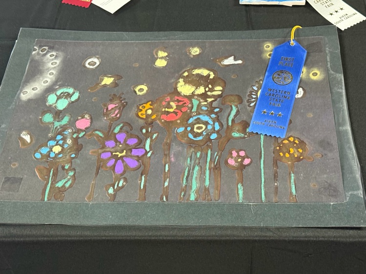 Art project on a black surface with flowers, stems, and leaves. Blue ribbon with text.