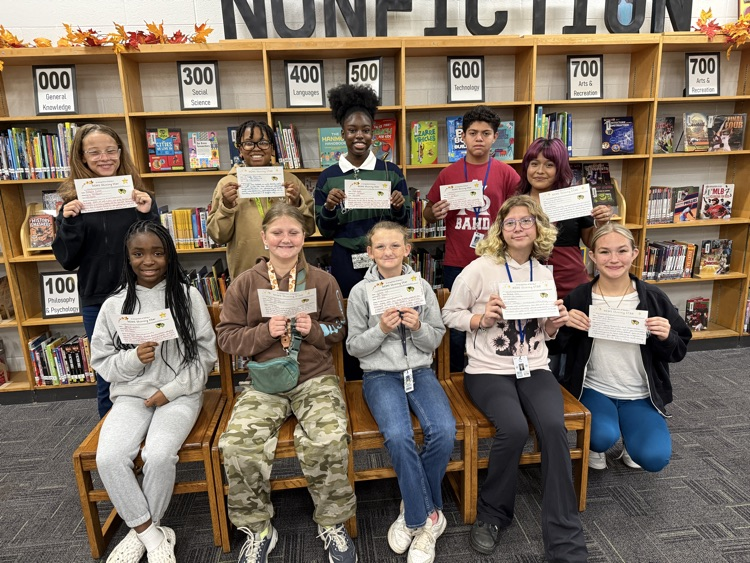 Students in a library pose with certificates, sitting on benches with bookshelves in the background.