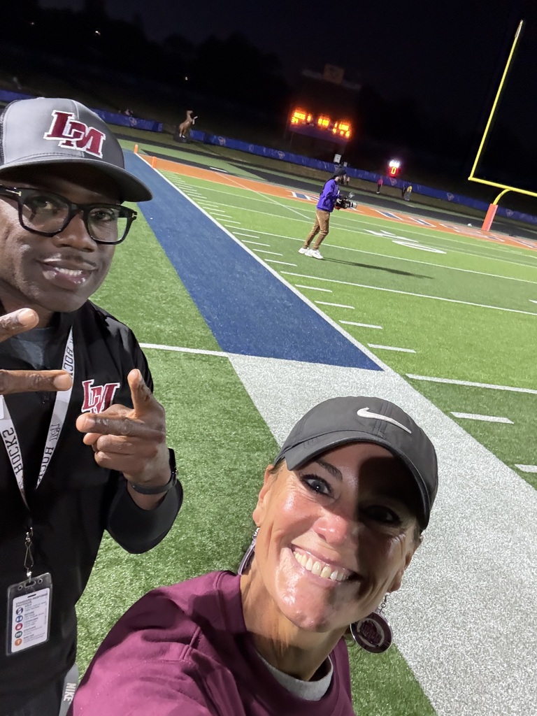 Two people stand on a football field at night. One person makes a peace sign, and the other smiles.