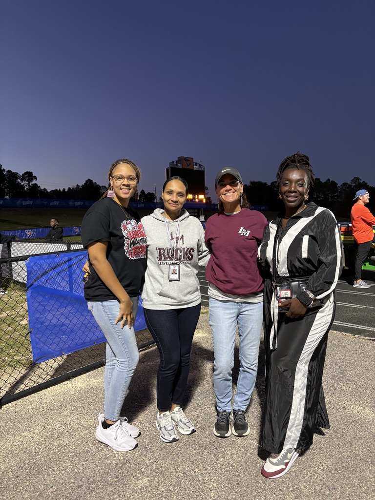 Four people stand in front of a fence at night. They wear casual clothes. A track is behind them.