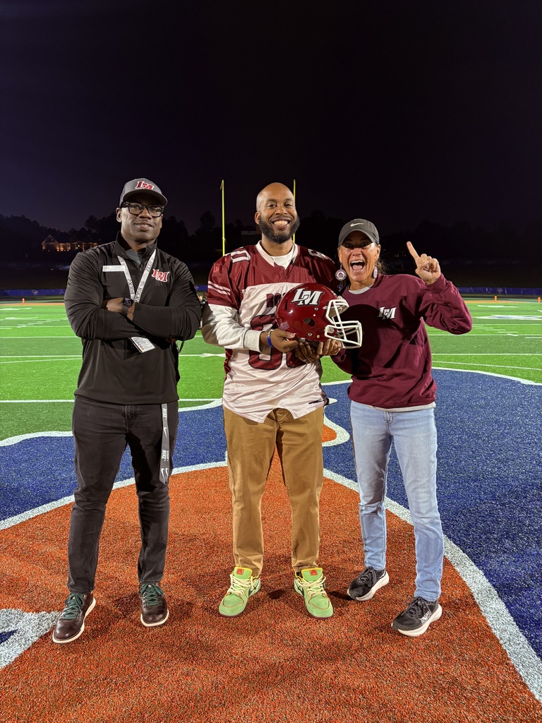 Three people on a football field at night, one holding a football helmet, with a field and scoreboard in the background.