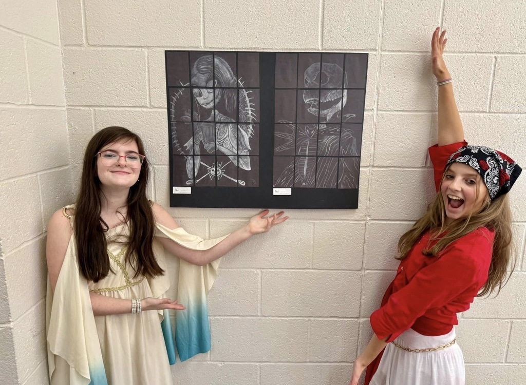 Two girls stand in a corner. One wears a costume, the other a red shirt and bandana. A framed picture hangs behind them.