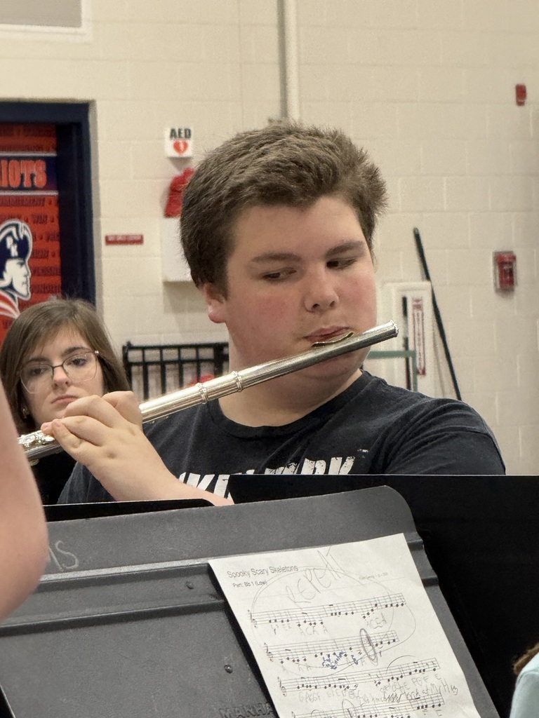 Boy plays flute while seated, looking at music sheet. Girl sits behind him, also playing flute.