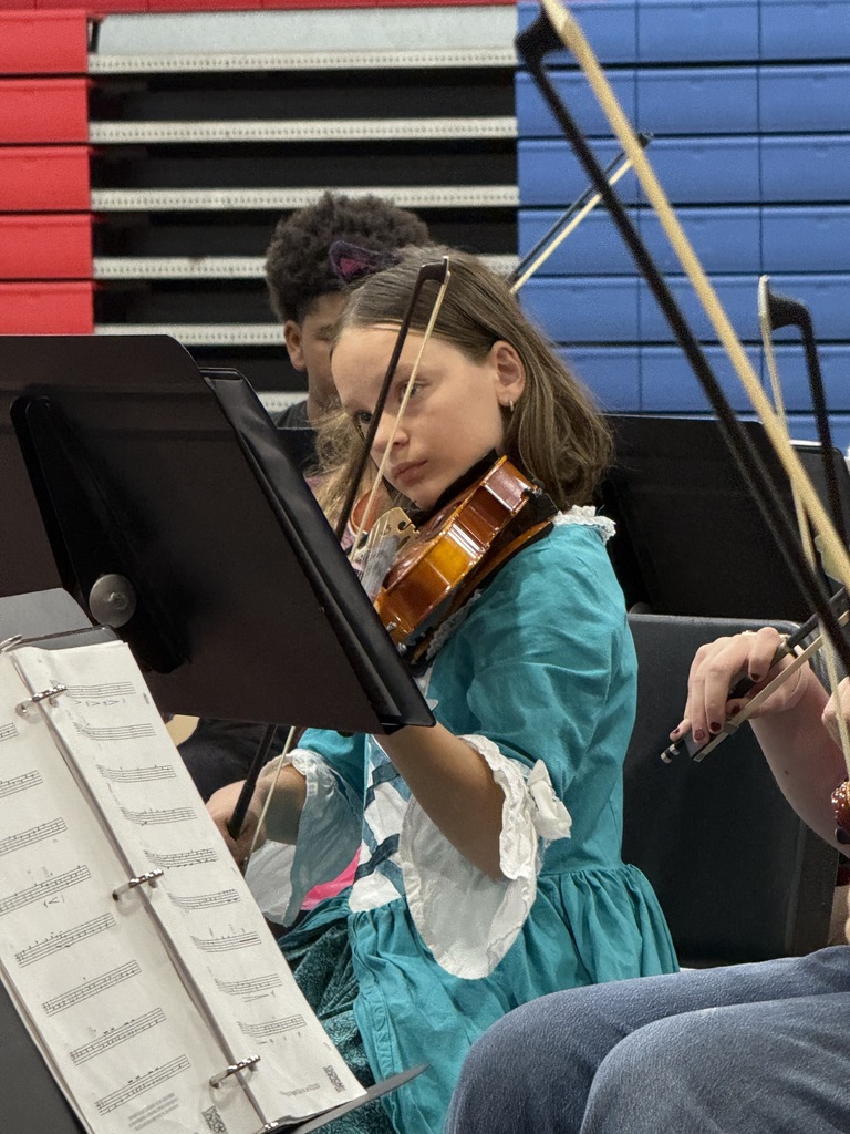 A young girl in a blue dress plays a violin while holding a bow in a classroom.
