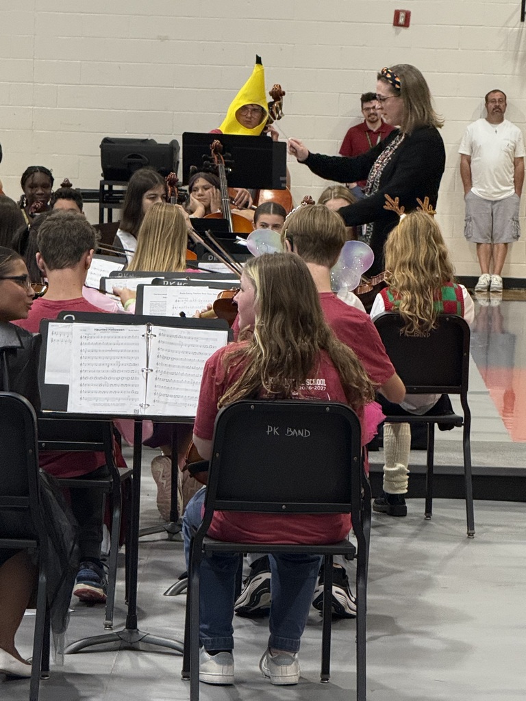 Students sit in chairs holding musical instruments; one woman in a red shirt directs while holding a music stand.