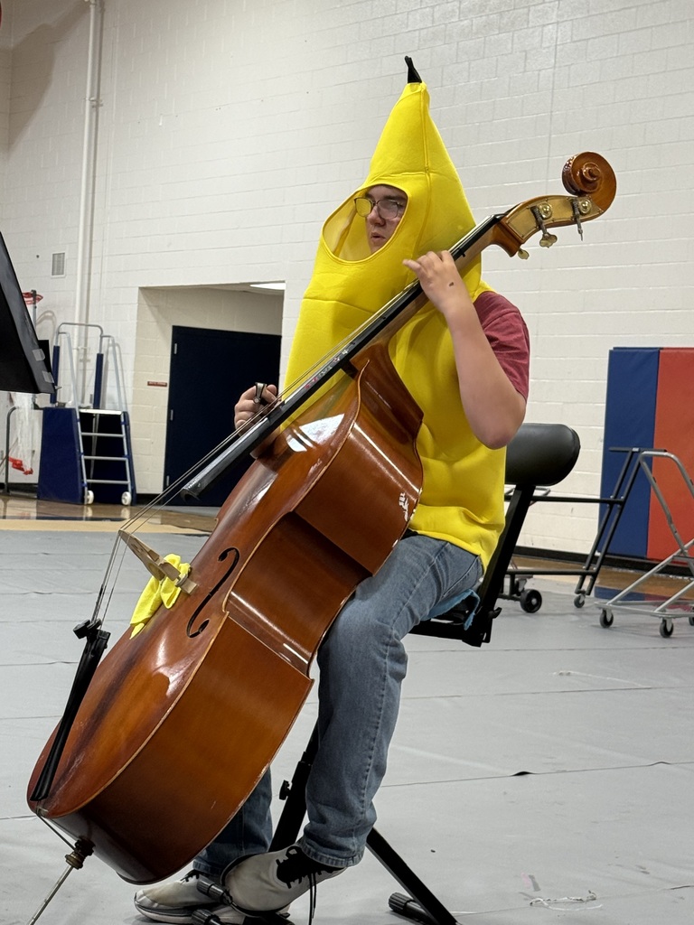 Person in banana costume seated on stool holding large cello, with music stand and doors in background.