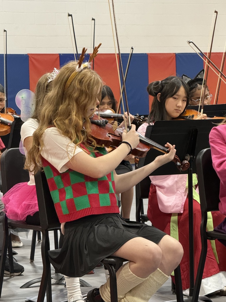 A student plays the violin in a large room with other students wearing Christmas costumes.