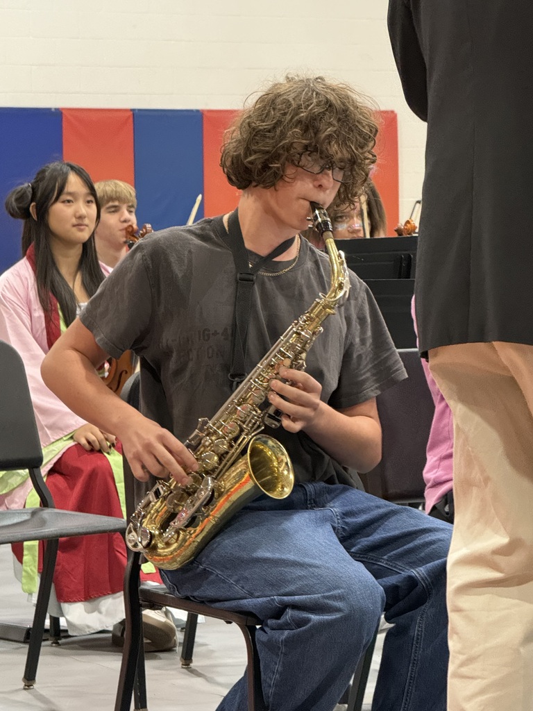 A seated boy plays a saxophone while other students watch. The boy has curly hair.