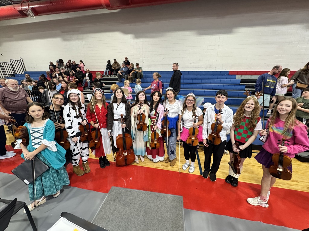 A group of students pose with string instruments in a gymnasium with a red and gray floor.