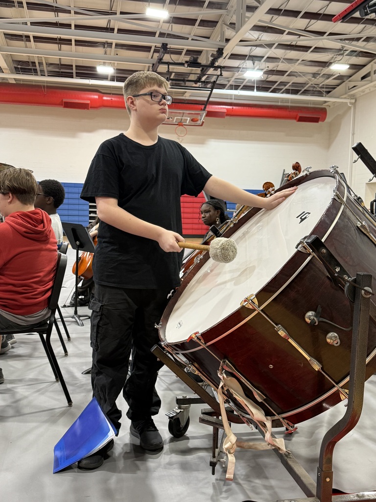 A boy in a black shirt plays a drum while others sit on chairs, watching.