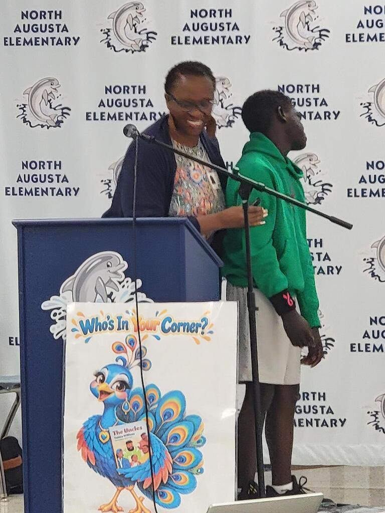 Two students at a podium with a microphone; one holds a sign reading "What's in the Corner?"