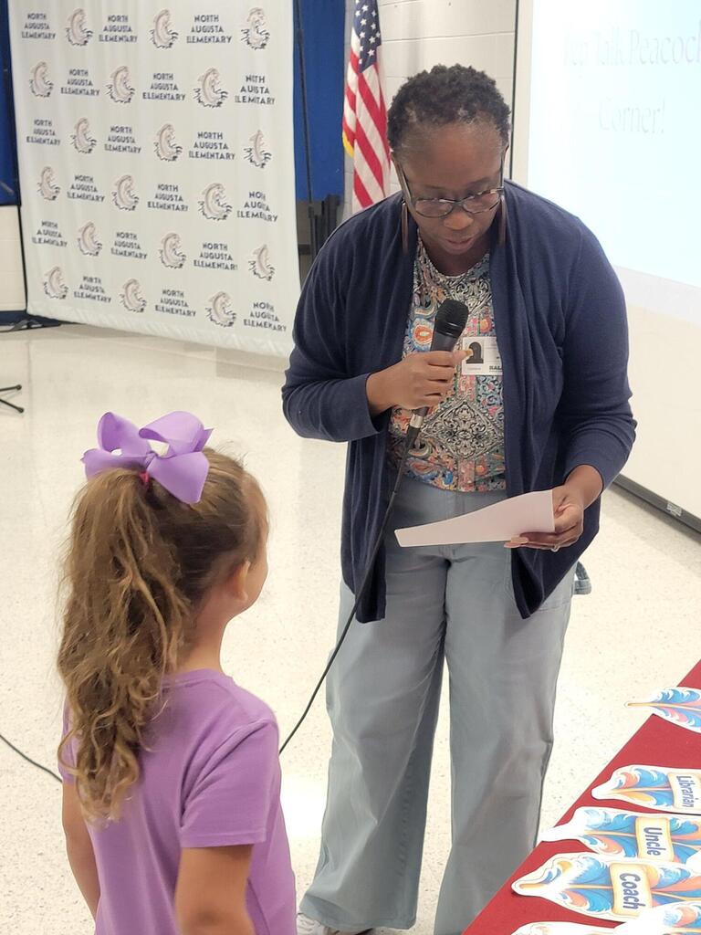 Woman with microphone and paper speaks to girl with purple bow; American flag and display board behind them.