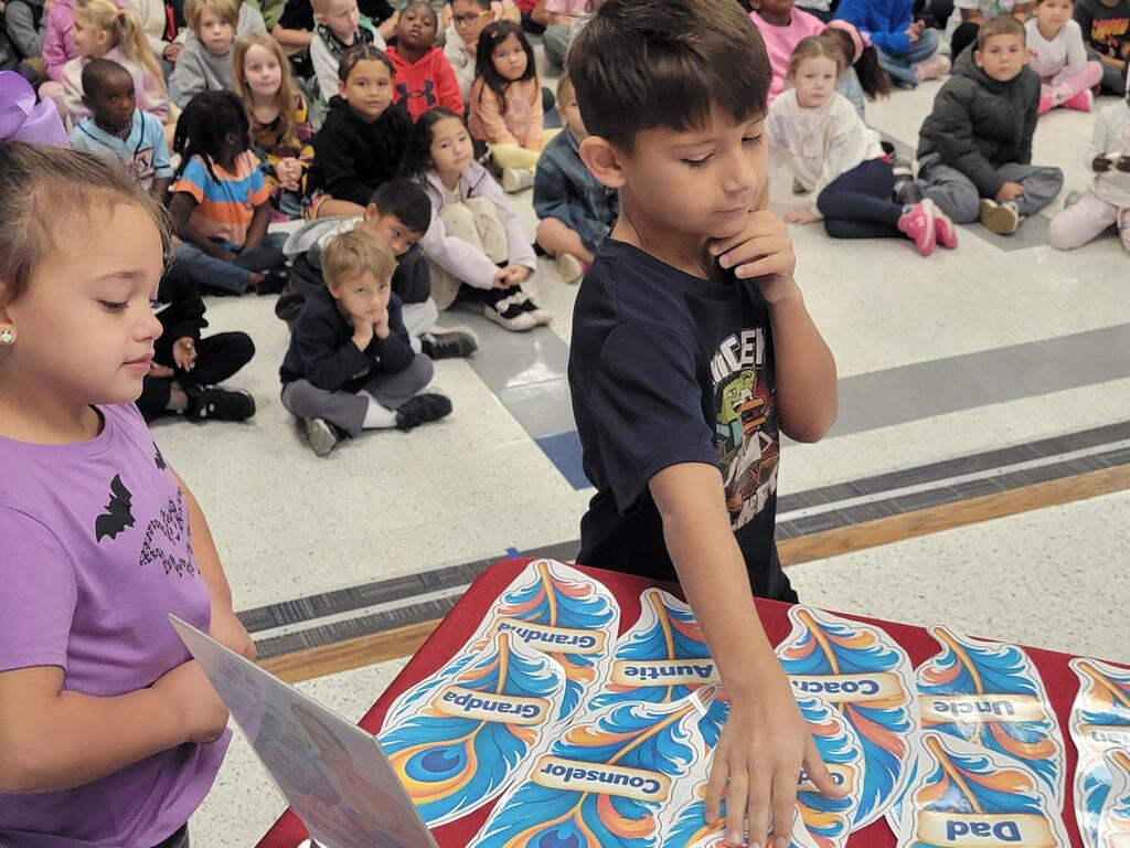 Children gathered in a classroom, one child looks at paper while another child touches a colorful table covering.