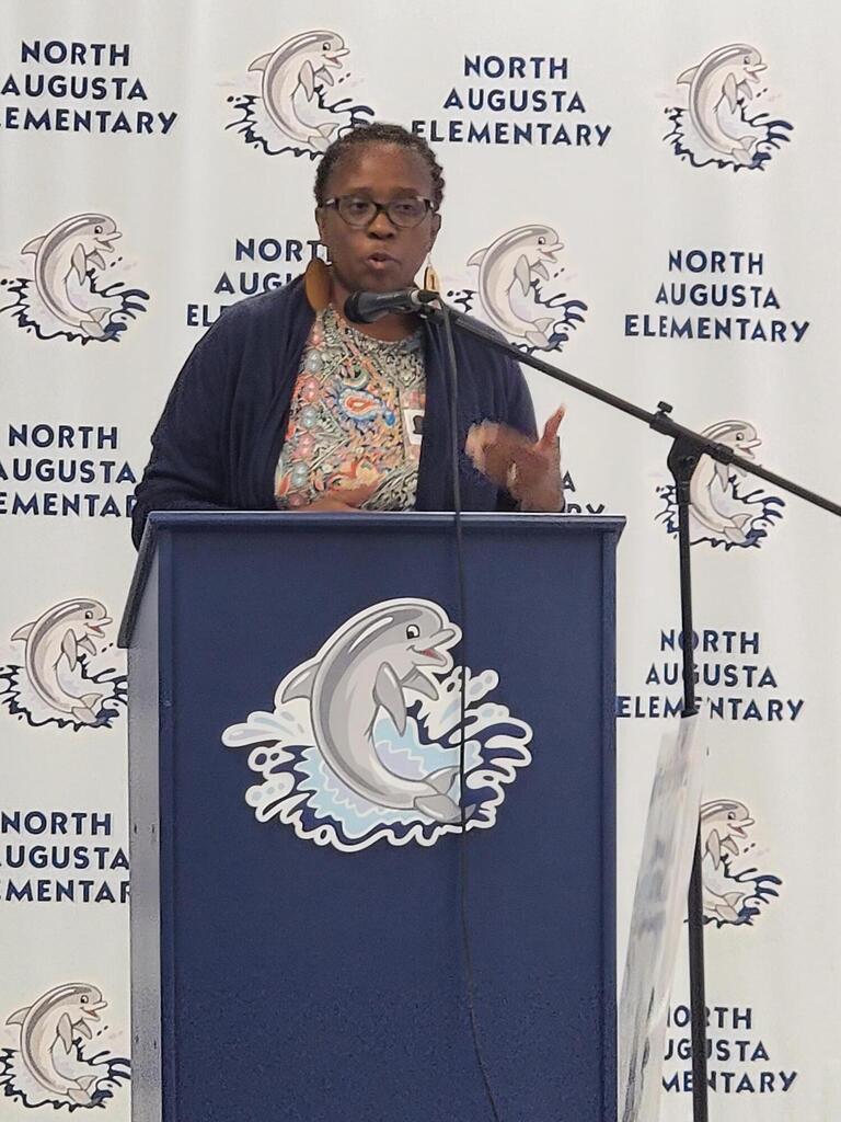 Woman speaking at podium with microphone, sign behind her reading North Augusta Elementary.