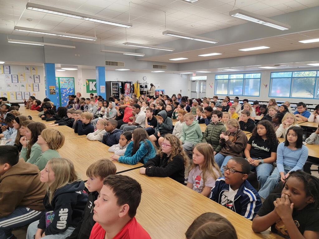 Many students sitting at long tables in a classroom, with overhead lights and large windows.