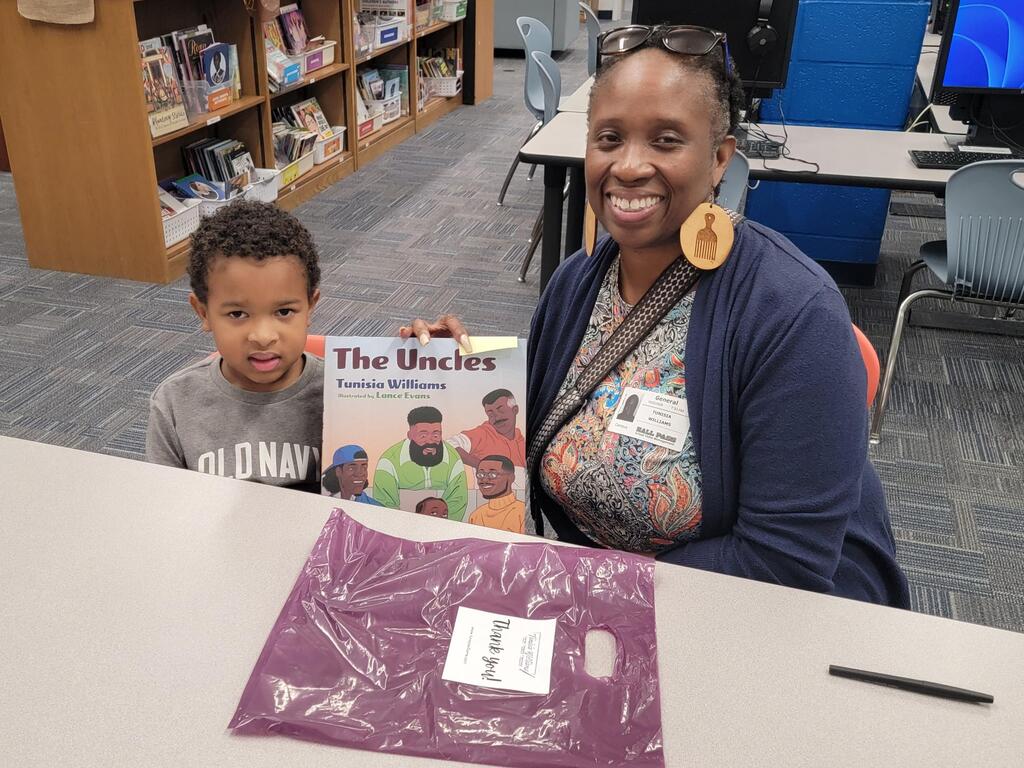 A woman and child sitting at a table, smiling. The child holds a book. Behind them, a bookshelf and desks.