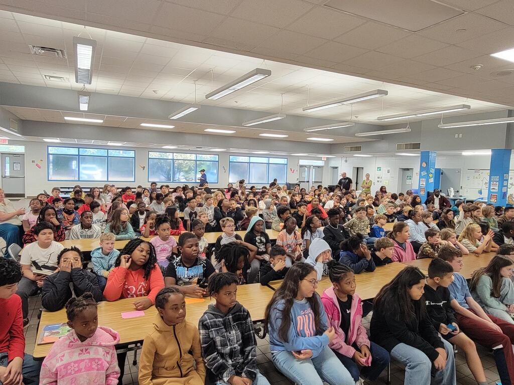 Large classroom with long tables, students sitting, some wearing jackets, and overhead lights.