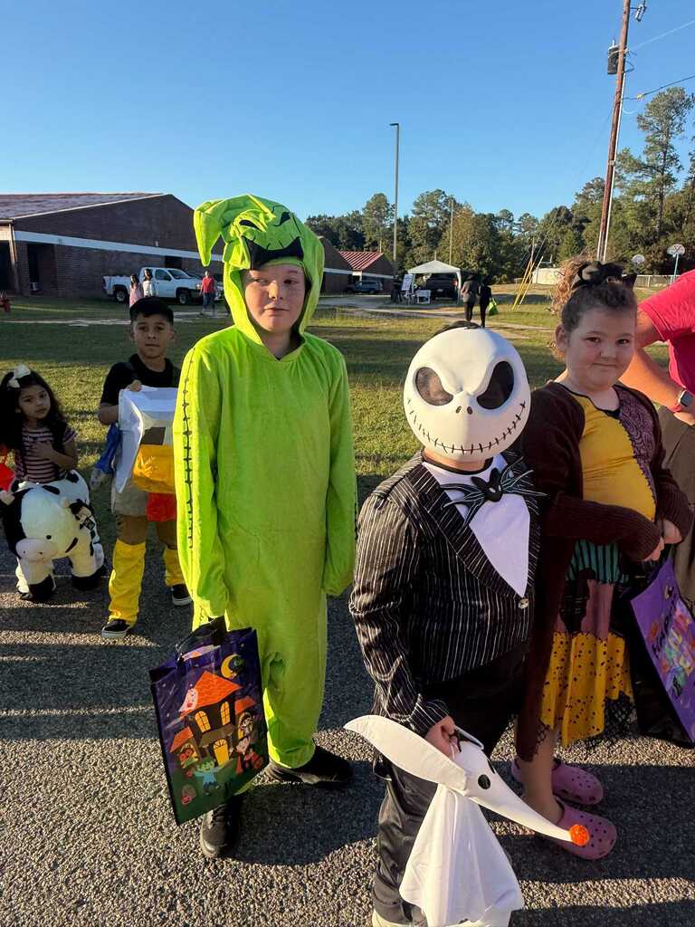 Five children in Halloween costumes, standing outdoors. One child in green is holding a bag.