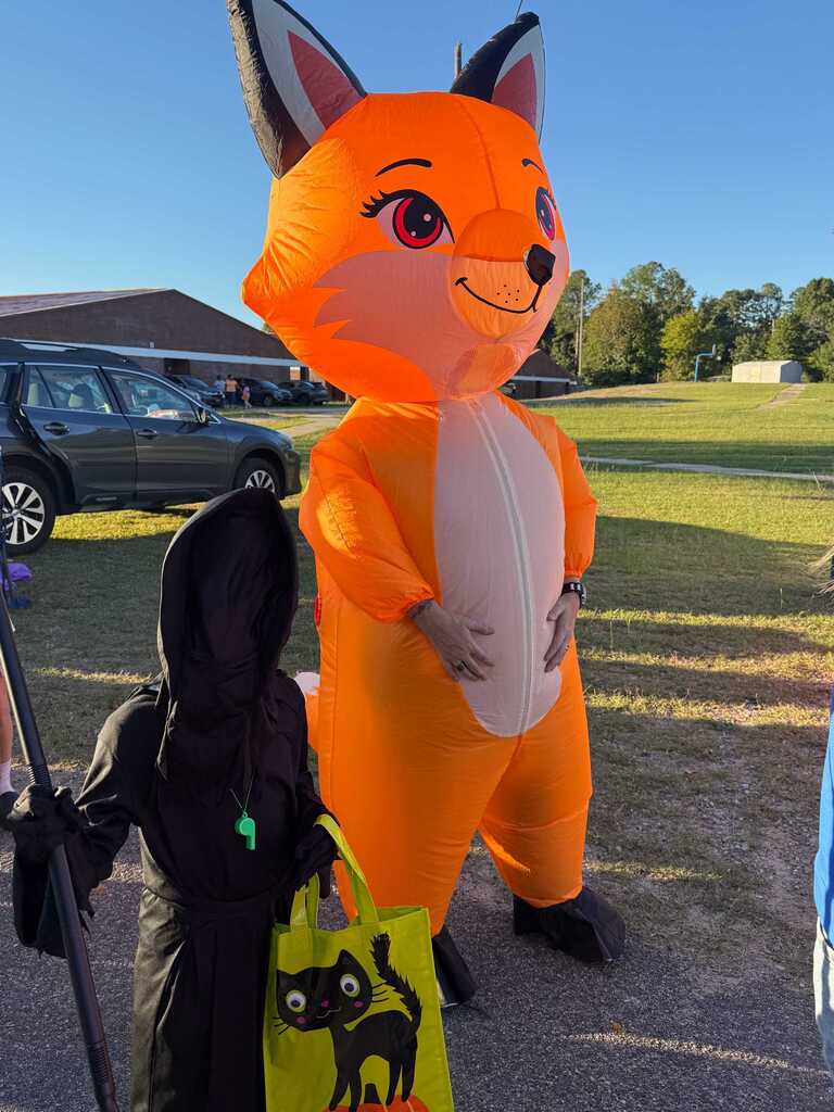 Person in costume holding a yellow bag with a cat picture stands next to large orange animal costume.