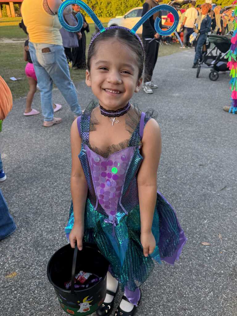 A child in a blue and purple costume holds a bucket of treats. Behind her, several people are walking.
