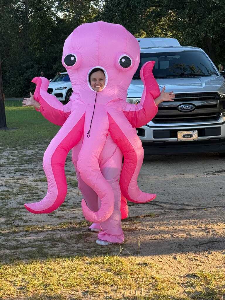 Person in pink octopus costume stands with arms outstretched in front of vehicle.