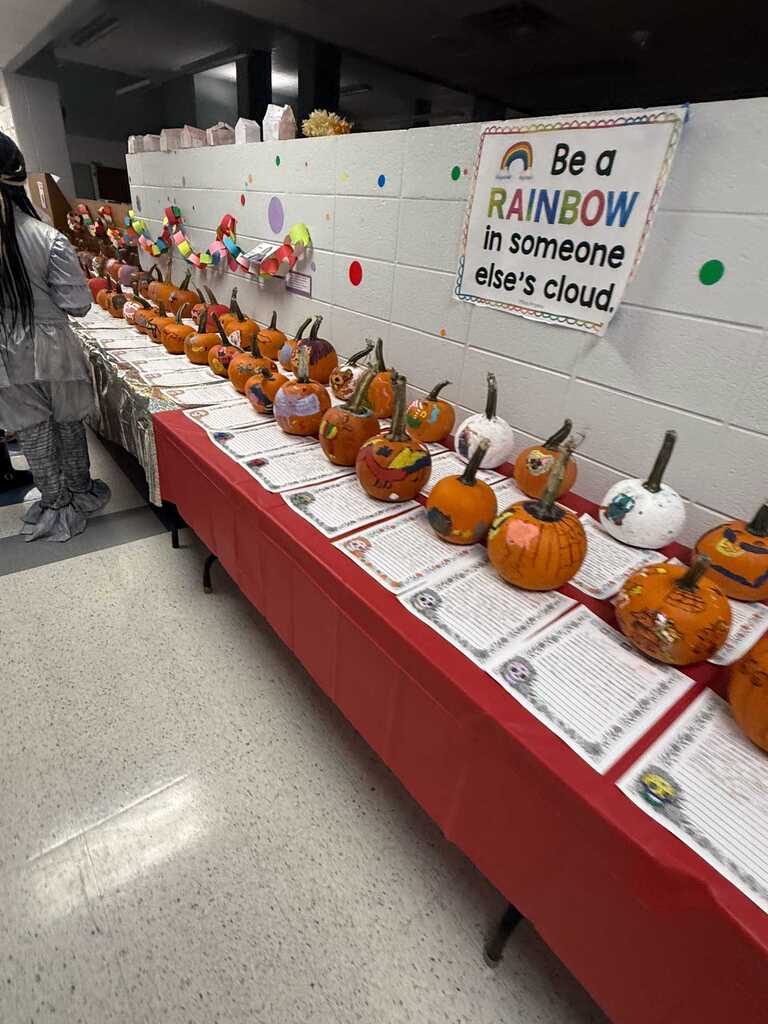 Long table with decorated pumpkins, papers, and a motivational sign on a wall in a classroom.