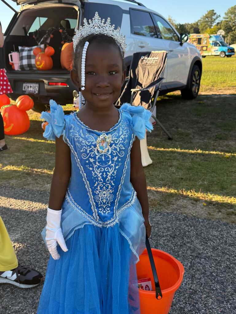 Child in blue dress with crown and white gloves holds orange bucket in parking lot with pumpkins.