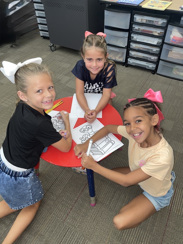 Three smiling girls are seated and coloring.