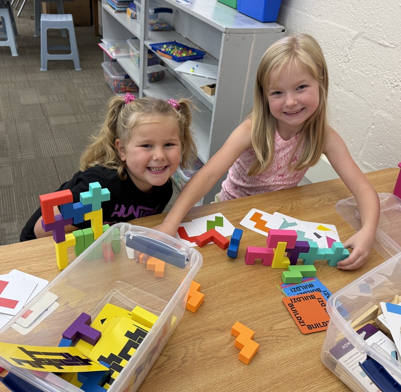 Two smiling girls sit at a table playing with blocks.