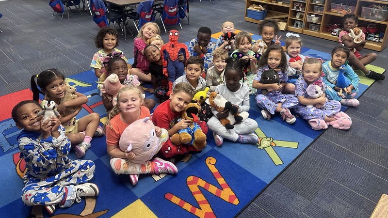 A class of kindergarten students sit on the floor wearing pajamas and holding stuffed animals.