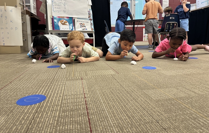 Four 4K students crawling on the floor are blowing cotton balls across the floor with straws.