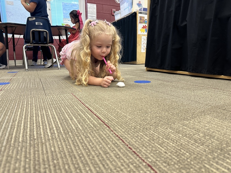 A girl is crawling on the floor is blowing cotton ball across the floor with a straw.