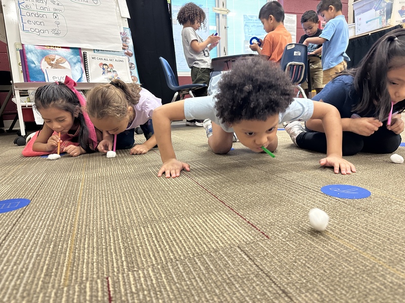 Four 4K students crawling on the floor are blowing cotton balls across the floor with straws.