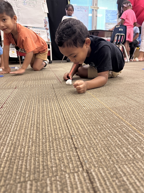A boy is crawling on the floor is blowing cotton ball across the floor with a straw.