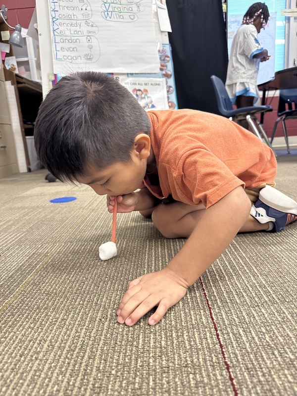 A boy is crawling on the floor is blowing cotton ball across the floor with a straw.
