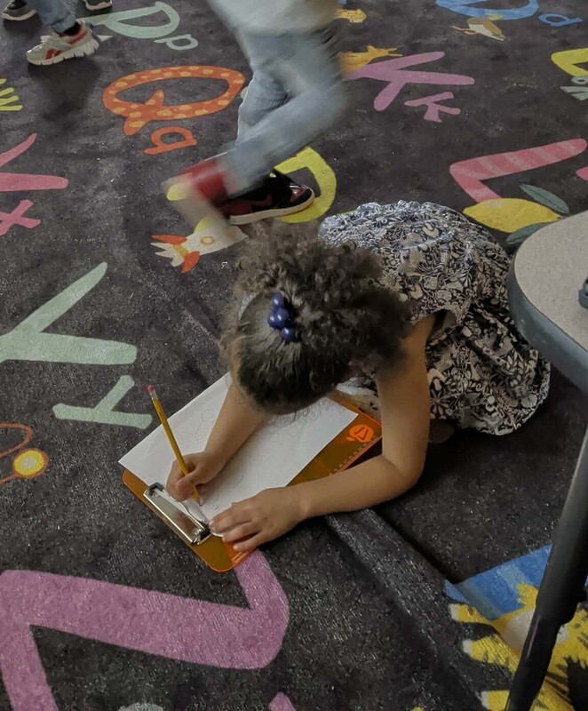 A girl is writing on a clpboard while sitting on an alphabet carpet.
