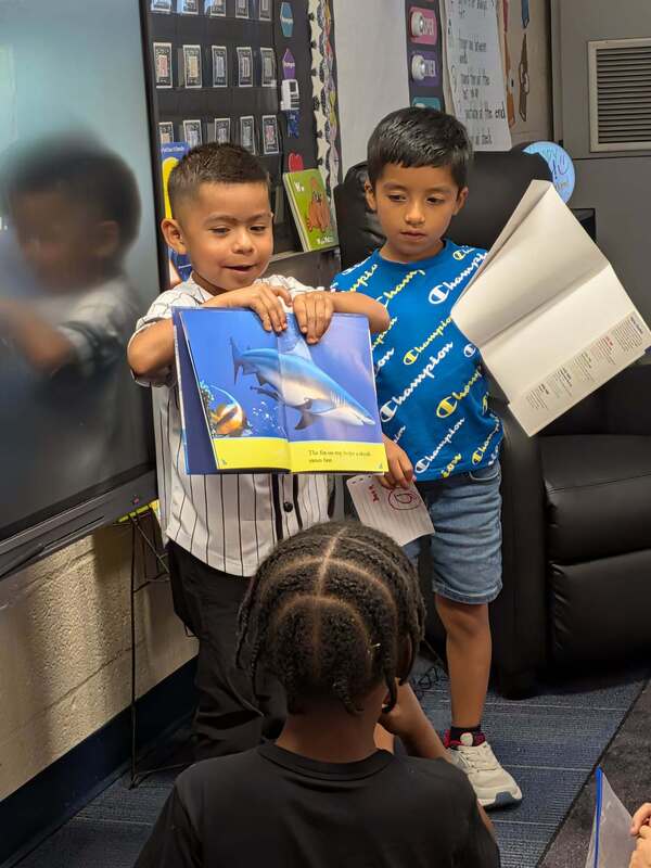 Two boys are holding up a book in front of their class.