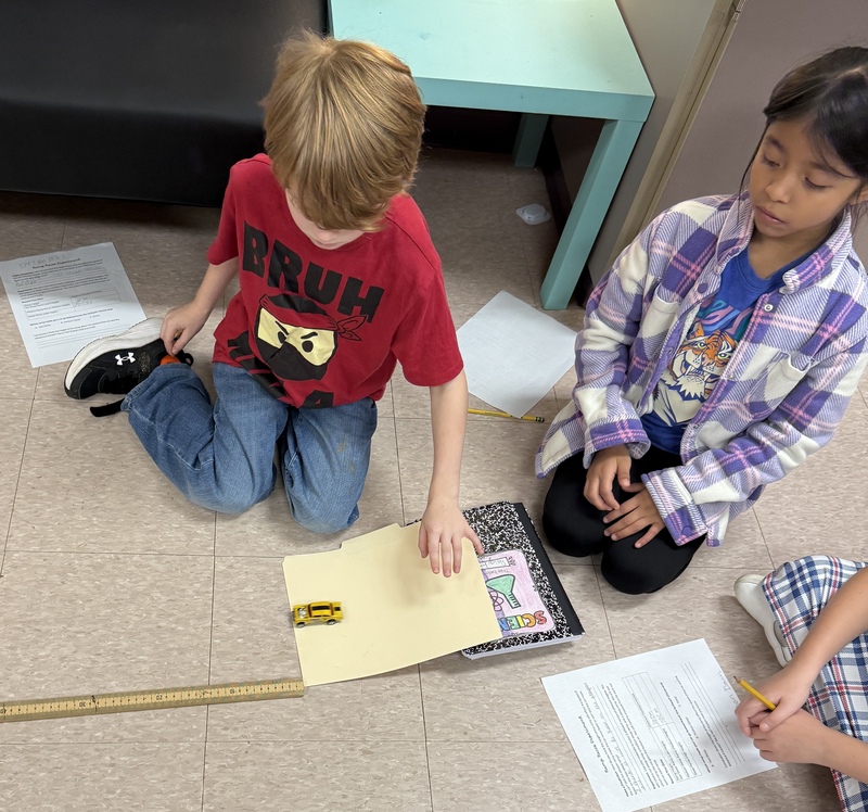Three students sit on the floor conducting a science experiment with a toy car.
