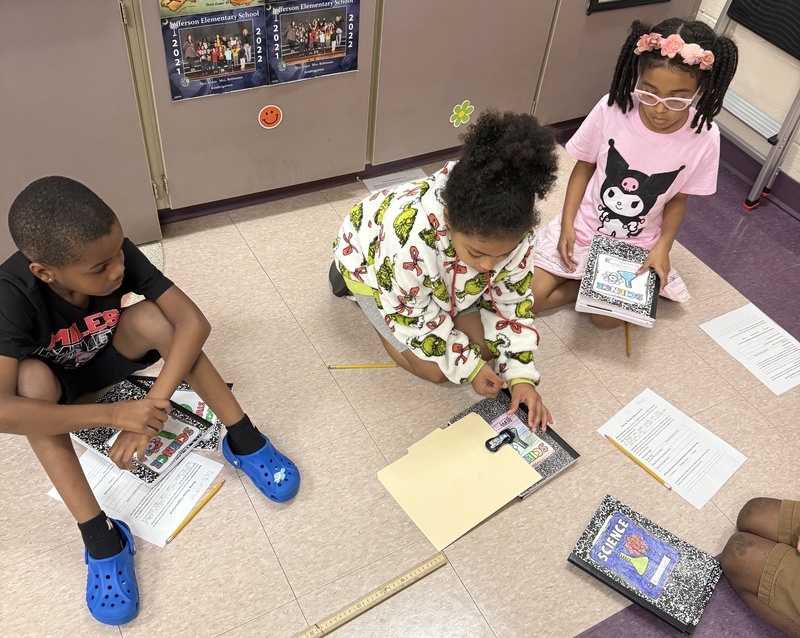 A group of students sit on the floor conducting a science experiment with a toy car.