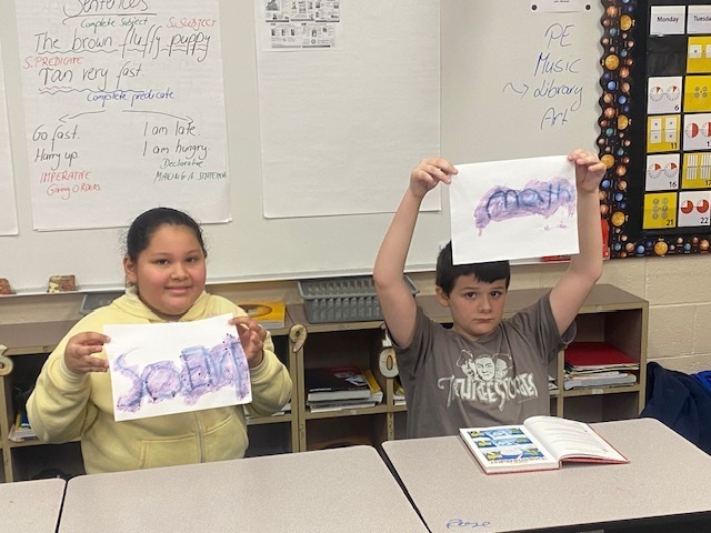 Two students hold up their sheets of paper to show their hidden messages.