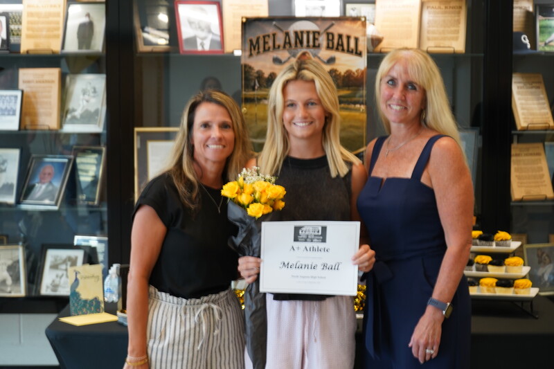 Student holding award certificate and flowers standing with two adult women in front of trophy case.